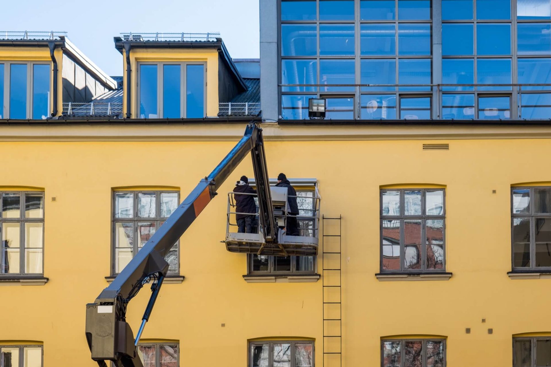 För att fräscha upp fasaden på en gul byggnad med stora fönster, använder arbetare en skylift i sitt arbete